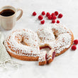 Kringle with white icing and red filling, accompanied by raspberries and a cup of coffee on a light surface.