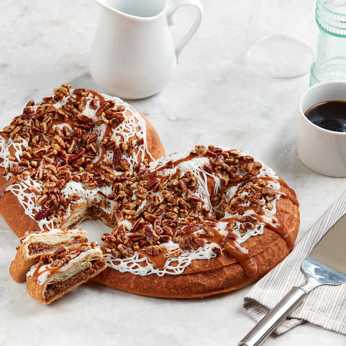 Pecan-topped kringle with a slice removed on a marble surface with a cup of coffee.